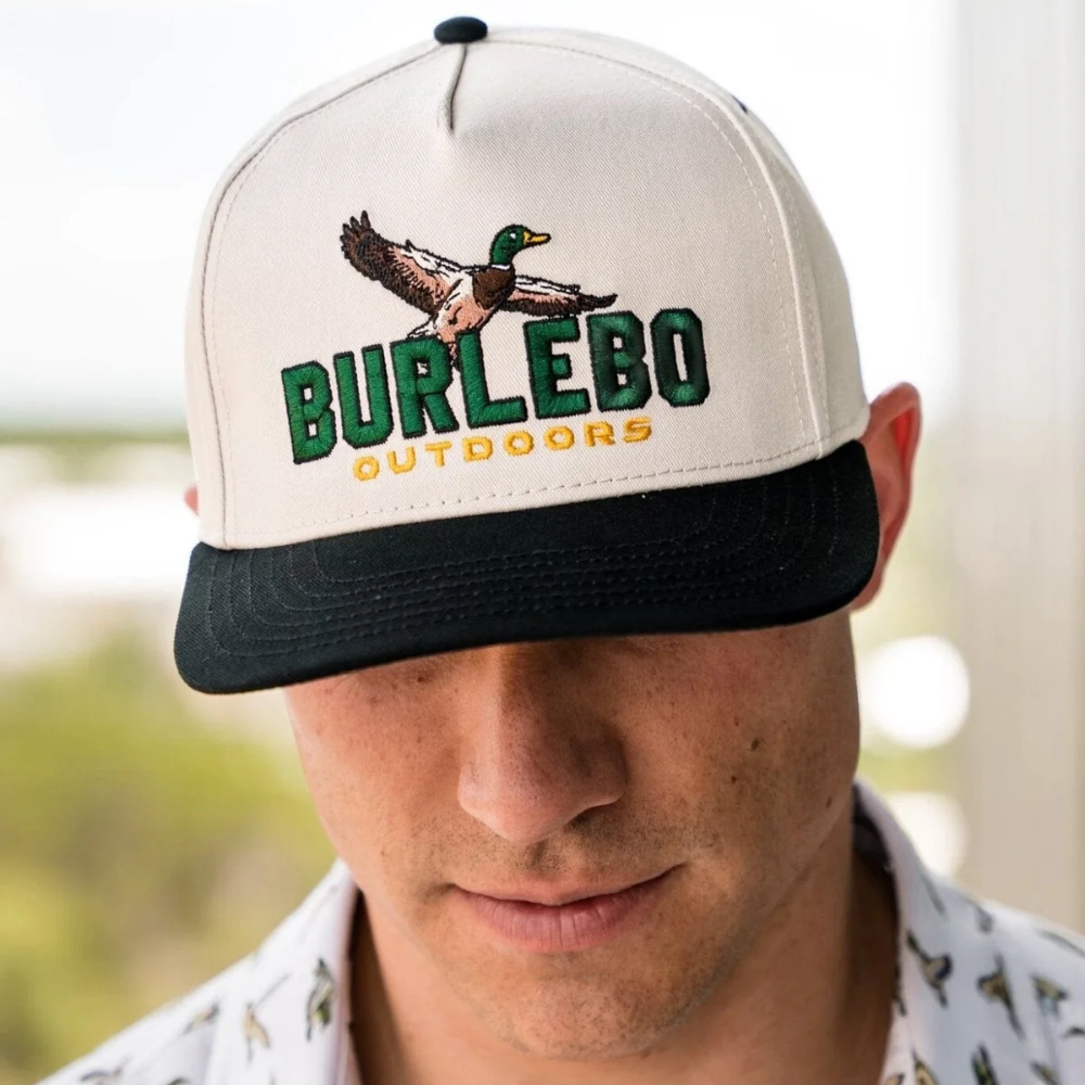 Mens snapback hat with a logo of a duck flying over a Burlebo Outdoors logo. The main part of the hat is the color cream, the bill and button on top of the hat are black. The Burlebo name is green, and the word outdoors is yellow.