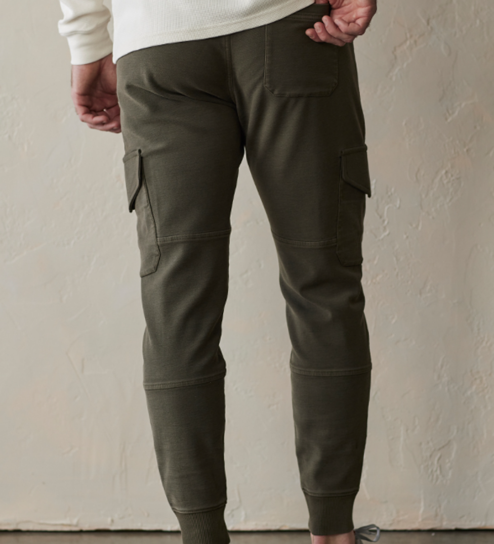 Back view of the Comfort Terry Jogger pants from The Normal Brand. Shown in color dusty olive green. The Jogger has draw strings at the waist, side pockets for your hands, and pockets on both legs, and a rear back pocket on the right side.