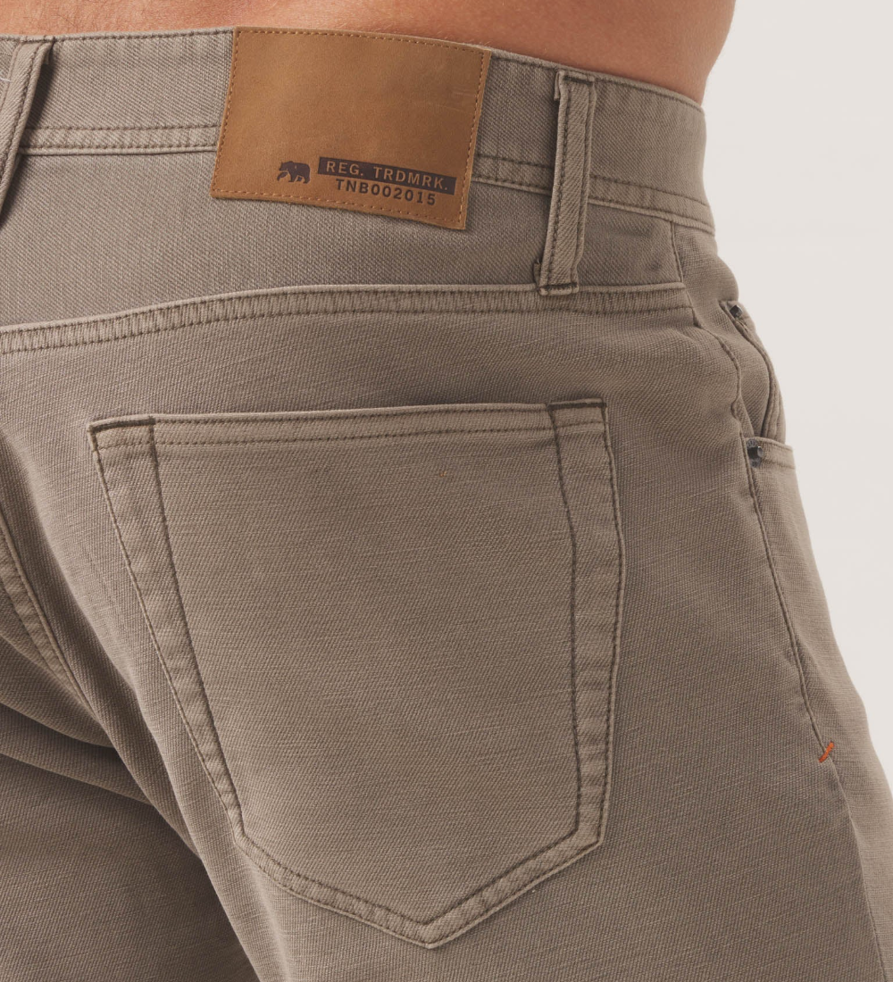Rear view of a pair of Comfort Terry Pant from The Normal Brand in the color taupe. Showing an up close view of the back right pocket, and a trademarked The Normal Brand logo.